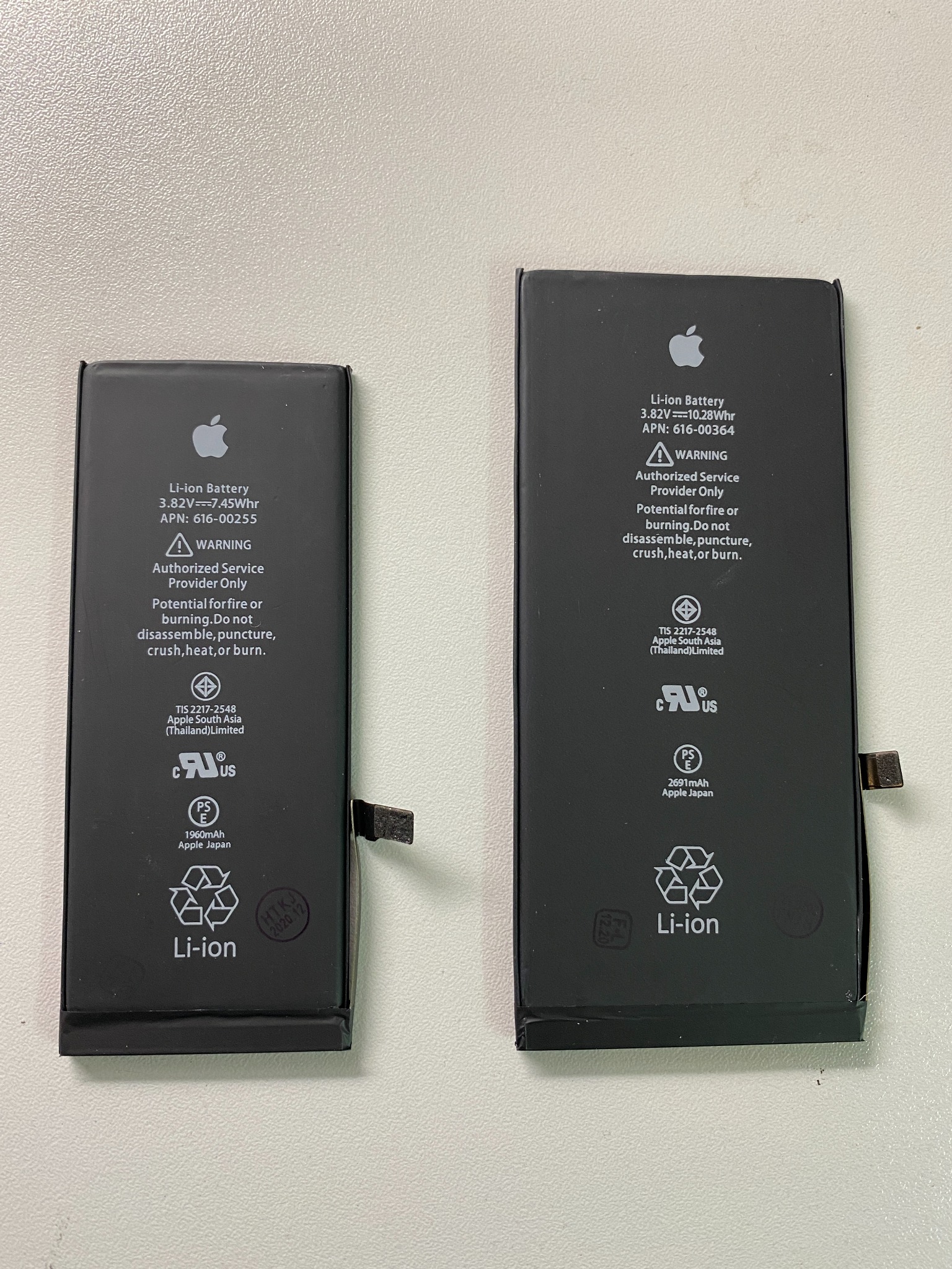 Dwie baterie litowo-jonowe do iPhone'a, jedna mniejsza i jedna większa, leżą obok siebie na białym tle w Poznaniu. Na każdej baterii widoczne ostrzeżenia i specyfikacje techniczne.