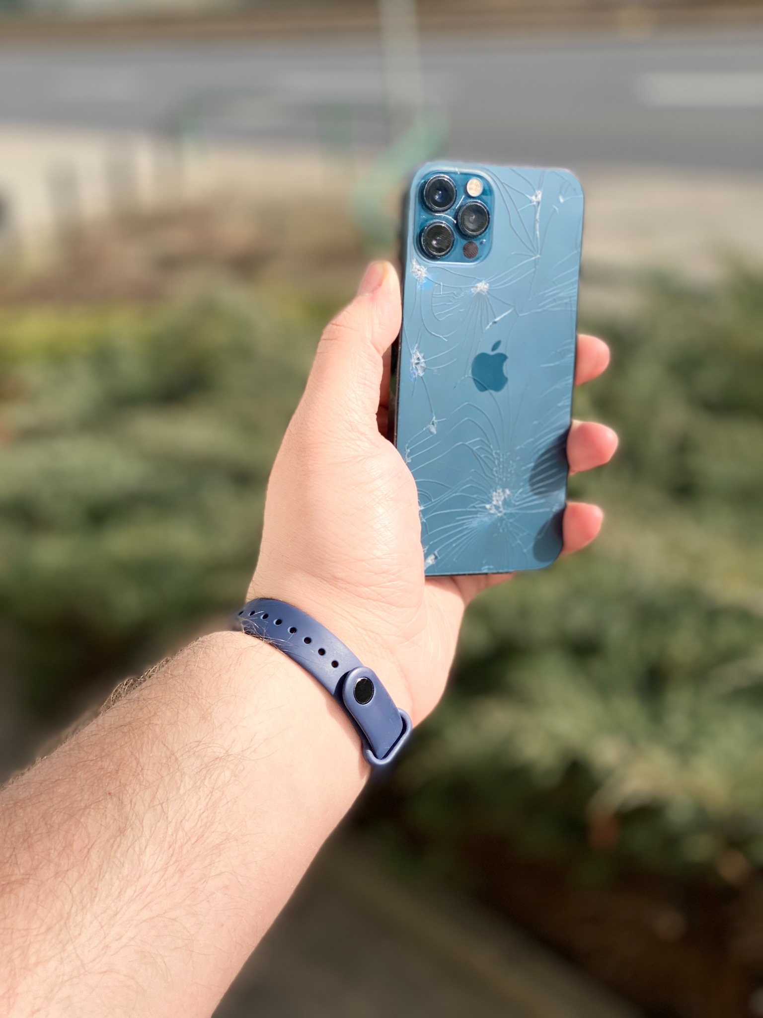 Mężczyzna trzyma w dłoni telefon iPhone z mocno pękniętą, niebieską obudową, widoczne liczne pęknięcia na całym tyle obudowy. Na nadgarstku widoczna niebieska opaska sportowa.