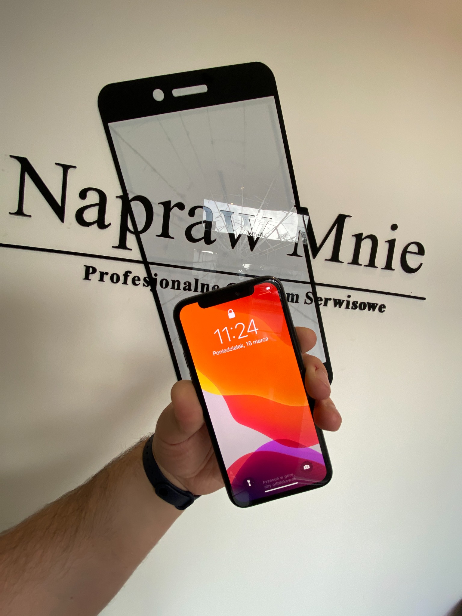 Smartfon iPhone z włączonym ekranem, trzymany w dłoni na tle szyldu 'Napraw Mnie' i przygotowanej do wymiany pękniętej szybki.