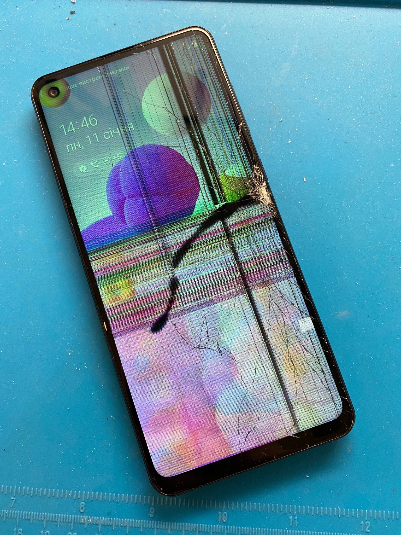 Smartfon z mocno uszkodzonym wyświetlaczem, widoczne pęknięcia i smugi na ekranie, leżący na niebieskiej macie serwisowej.
