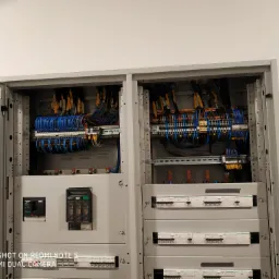 Otwarta szafa elektryczna z widocznymi przewodami, bezpiecznikami i elementami sterującymi; przewody uporządkowane w wiązki, oznacznikami w kolorze żółtym, przewody w kolorze niebieskim i brązowym;...