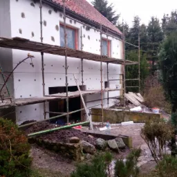 Dom w trakcie ocieplania, widoczne rusztowania, styropian na ścianach z zaślepkami, przygotowane okna, teren wokół z elementami ogrodu.