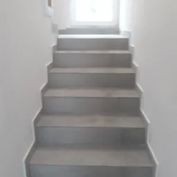 Schody wyłożone szarymi płytkami ceramicznymi, widok z dołu, białe ściany i okno w tle.