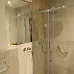 Nowoczesna kabina prysznicowa z deszczownicą i uchwytem dla osób niepełnosprawnych, widoczny fragment toalety i umywalki, jasne płytki imitujące drewno na podłodze.