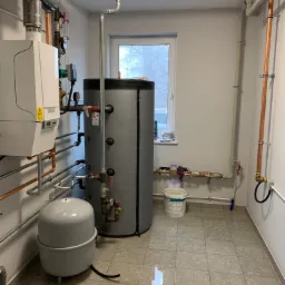 Kotłownia z piecem gazowym, zasobnikiem ciepłej wody, naczyniem przeponowym i instalacją hydrauliczną z miedzianych rur, widok na okno z parapetem.