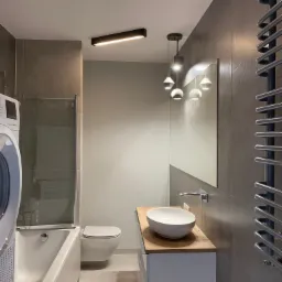 Nowoczesna łazienka z szarymi płytkami, białym umywalką na drewnianym blacie, podwieszaną toaletą i grzejnikiem drabinkowym, oświetlona podłużnymi lampami sufitowymi i wiszącymi lampami kulami.