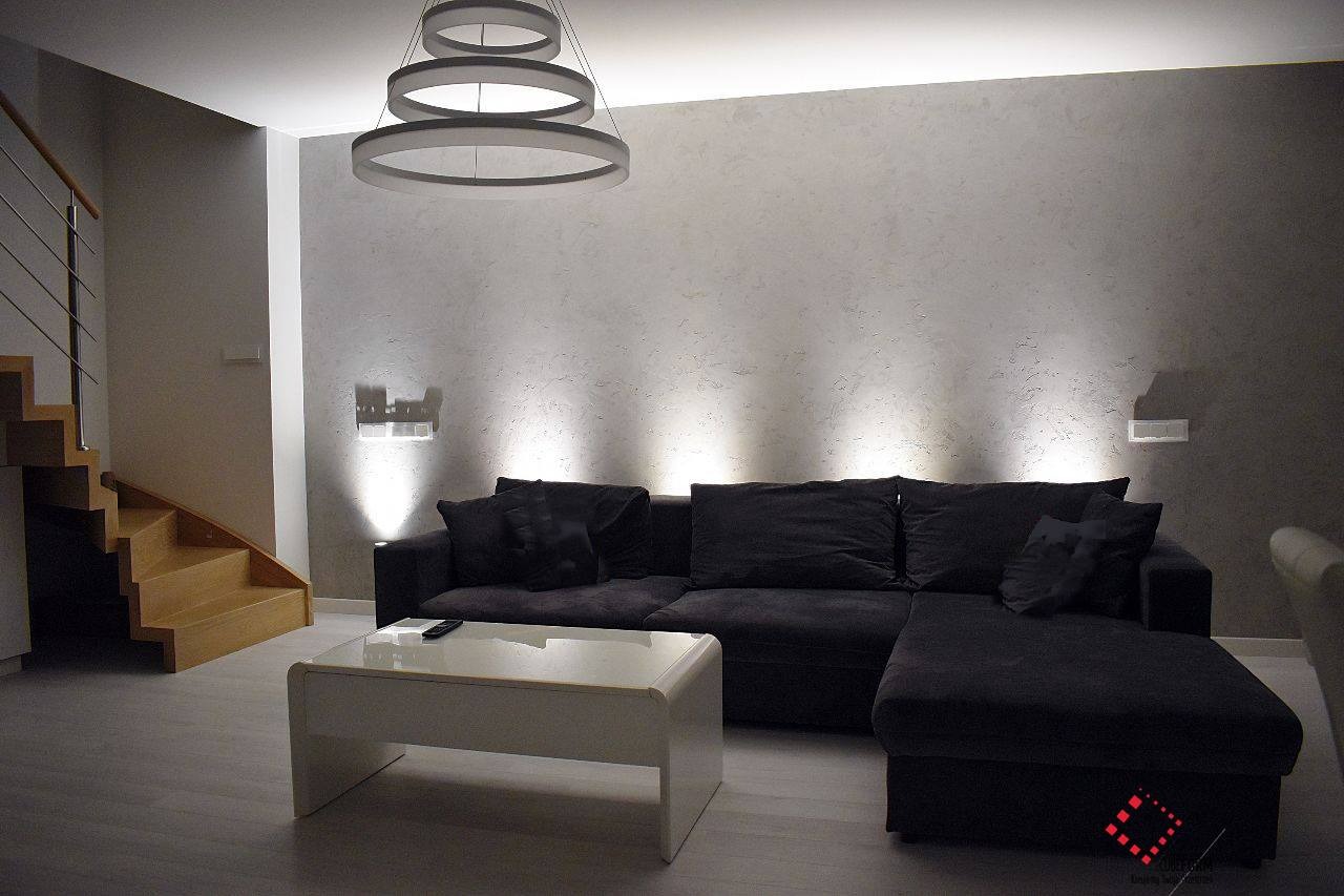 Nowoczesny salon z narożną sofą w odcieniach szarości, drewnianymi schodami z metalową balustradą i designerską lampą pierścieniową podwieszoną pod sufitem. Na ścianie dekoracyjne oświetlenie...