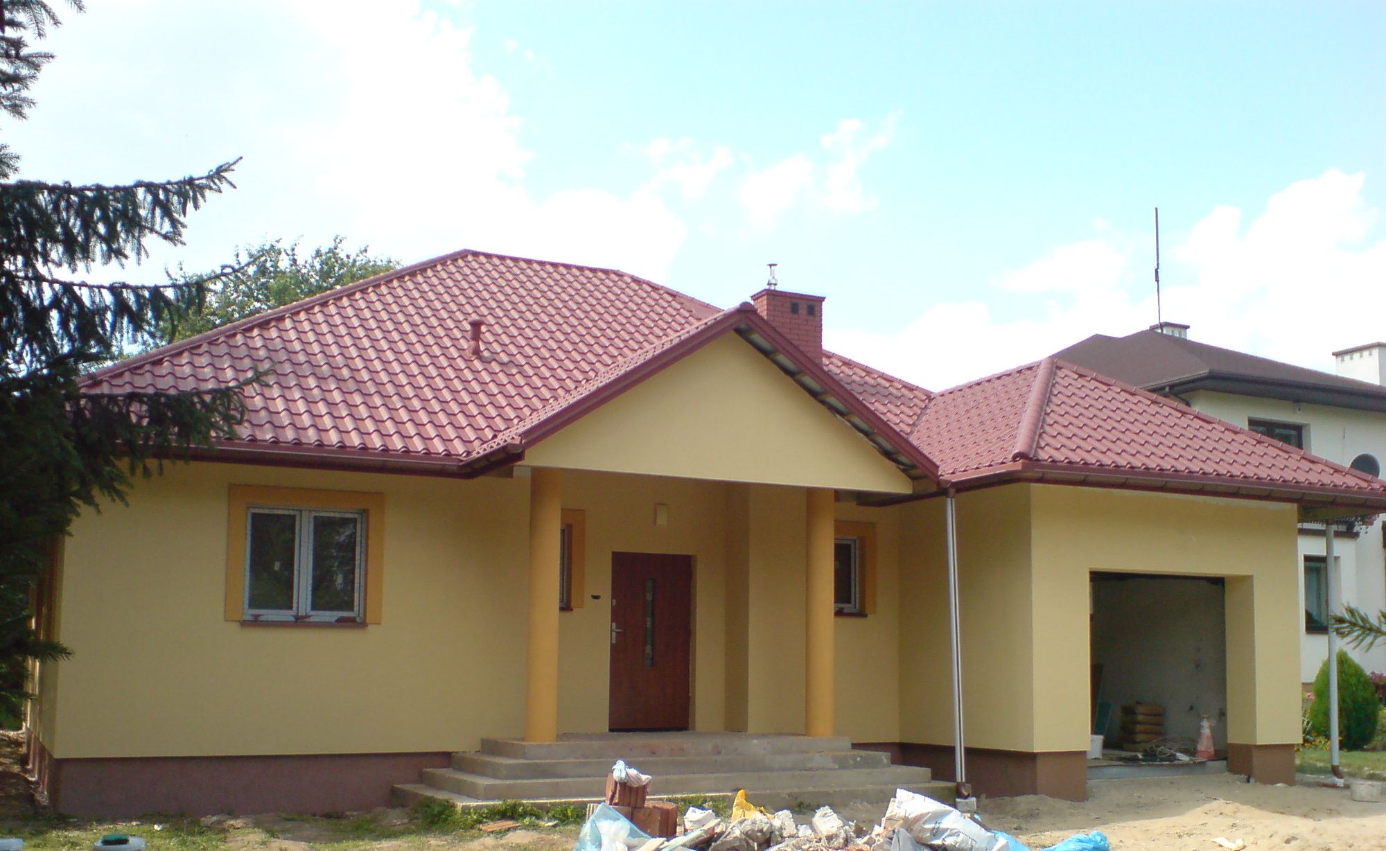 Żółty dom jednorodzinny z czerwoną dachówką, widoczne ślady prac budowlanych przed wejściem i w otwartym garażu.