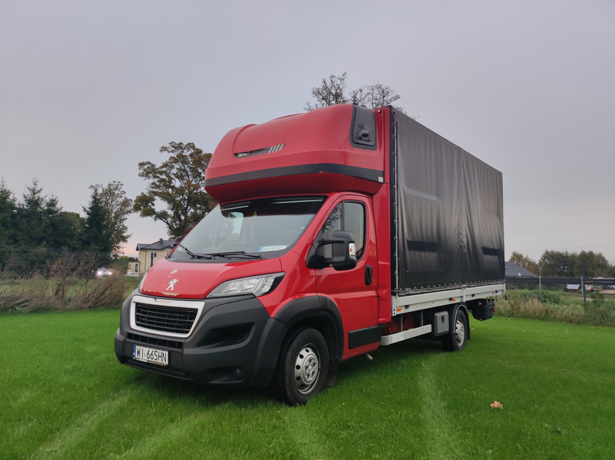 Czerwony samochód dostawczy Peugeot z plandeką na zielonej trawie, widok z przodu, tablice rejestracyjne WI 665HN.