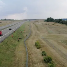 Tractor with a rotary mower mowing grass on a large, grassy area next to a highway, visible workers in orange vests along the fence.