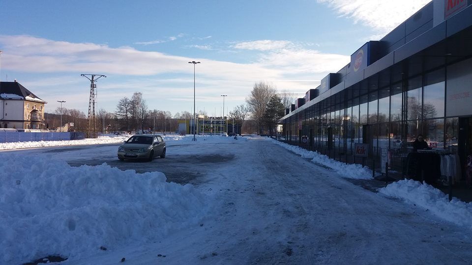 Zimowy krajobraz parkingu przed centrum handlowym z odśnieżoną nawierzchnią i stertami śniegu po bokach, zaparkowany samochód osobowy w oddali.