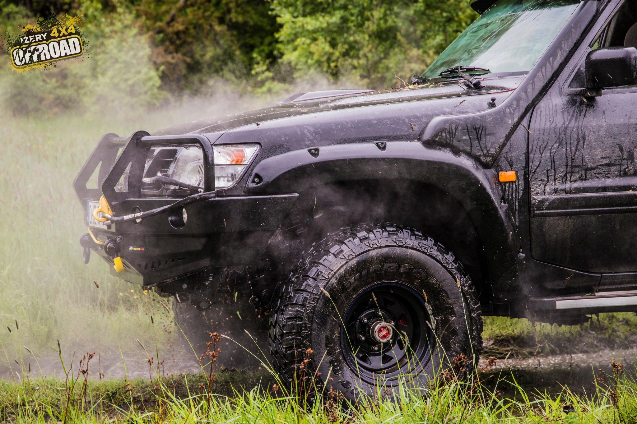 Czarny, błotnisty samochód terenowy 4x4 z logiem na szybie, w trakcie jazdy po trawiastym terenie, unoszący tumany pary i błota.