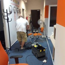 Czysta Panda Pranie Tapicerki Meblowej i Wykładzin - Pracownik w białej koszulce i beżowych spodniach czyści niebieską wykładzinę w biurze za pomocą szarego odkurzacza przemysłowego marki Karcher, w tle pomarańczowe krzesła biurowe i ściana...