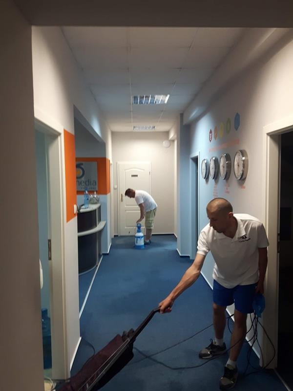 Pracownik w białej koszulce i niebieskich spodenkach odkurza granatowy dywan w korytarzu biurowym, w tle drugi pracownik używa spryskiwacza na dywanie, na ścianie widoczne zegary wskazujące różne...