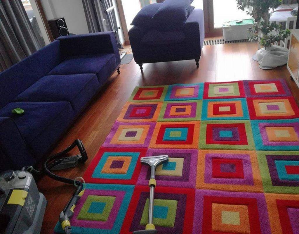 Wielobarwny dywan w kwadraty czyszczony odkurzaczem piorącym, widoczny fragment sofy i fotela w tle.