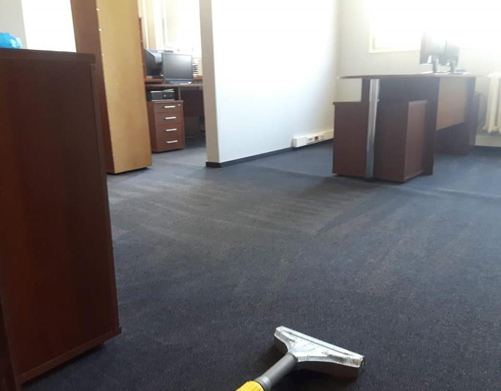 Fragment biura z ciemną wykładziną dywanową, na której widoczne są ślady po czyszczeniu, z dyszą odkurzacza piorącego w dolnej części kadru.