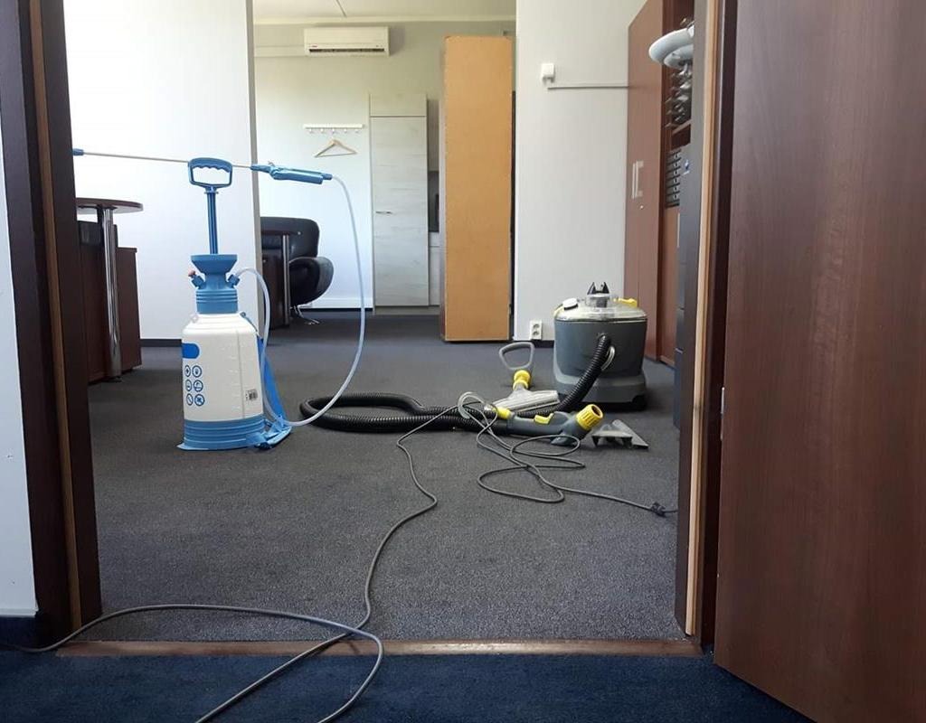 Sprzęt do czyszczenia wykładzin biurowych: spryskiwacz ciśnieniowy z lancą, odkurzacz piorący z końcówkami, rozłożone na szarej wykładzinie w pomieszczeniu biurowym.