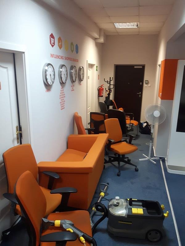 Wnętrze biura z niebieską wykładziną i pomarańczowymi meblami przygotowywane do czyszczenia tapicerki, widoczny sprzęt do prania ekstrakcyjnego, krzesła i sofa przesunięte na bok.