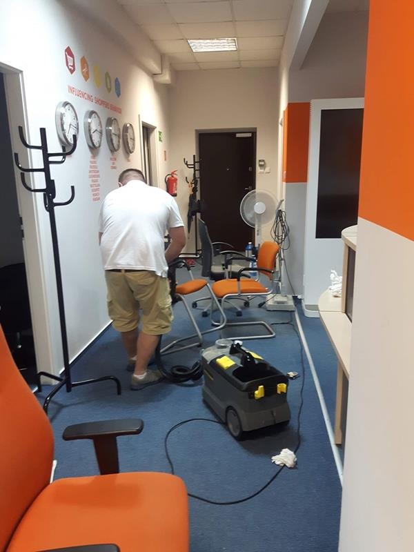 Pracownik w białej koszulce i beżowych spodniach czyści niebieską wykładzinę w biurze za pomocą szarego odkurzacza przemysłowego marki Karcher, w tle pomarańczowe krzesła biurowe i ściana...