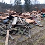 Stos gruzu i resztek drewnianych desek po rozbiórce budynku, widoczne ślady opon na trawie, w tle drzewa.
