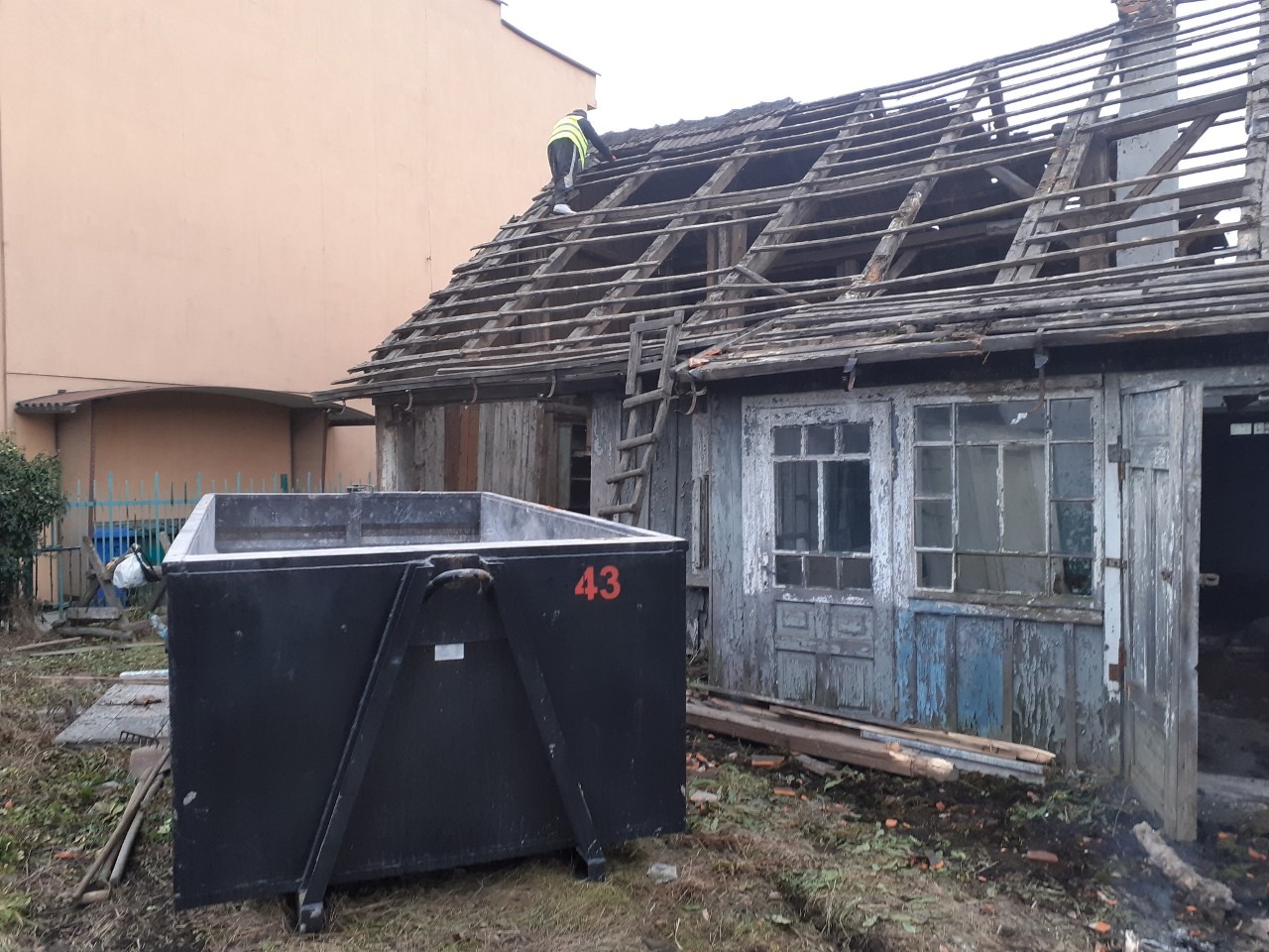 Demontaż dachu starego, drewnianego budynku. Widoczny pracownik w kamizelce na dachu, kontener na gruz z numerem 43, oraz zniszczona elewacja budynku z odpadającą farbą.