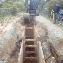 Głęboki wykop zabezpieczony szalunkiem stalowym, z widocznymi rurami w ziemi i koparką pracującą w tle, nadzorowany przez osobę w kamuflażu.