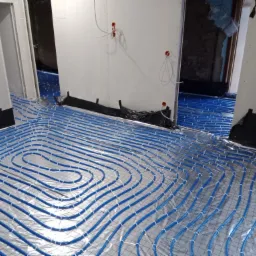 Niebieskie rury ogrzewania podłogowego ułożone na folii termoizolacyjnej w nowo wykończonym pomieszczeniu z widocznymi przewodami elektrycznymi.