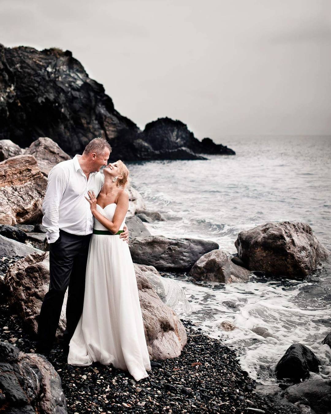 Para w strojach ślubnych stoi na skalistej plaży, obejmując się i patrząc sobie w oczy, z ciemnymi skałami i morzem w tle.