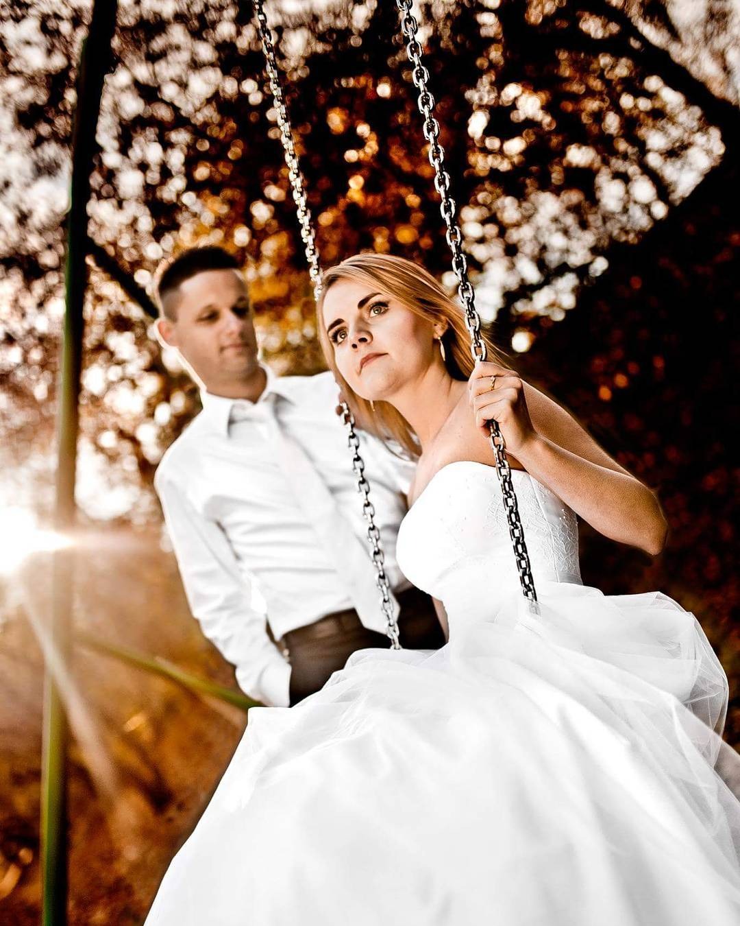 Para młoda na huśtawce w plenerze, panna młoda w białej sukni bez ramiączek, pan młody w białej koszuli, promienie słońca w tle.