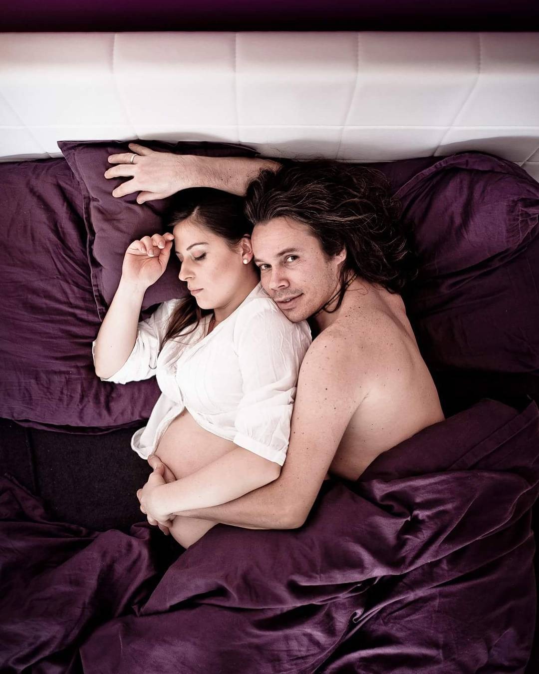 Para w łóżku; kobieta w białej koszuli, z widocznym ciążowym brzuchem, śpi, mężczyzna z długimi włosami obejmuje ją, patrząc w obiektyw, fioletowa pościel.