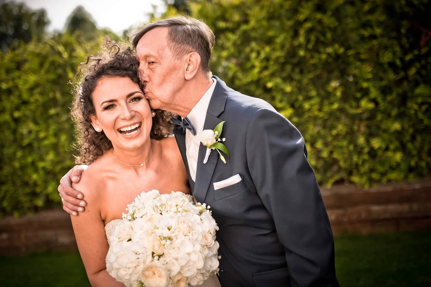 Uśmiechnięta panna młoda z kręconymi włosami, trzymająca bukiet białych róż, całowana w policzek przez elegancko ubranego mężczyznę w garniturze z muszką na tle zieleni.