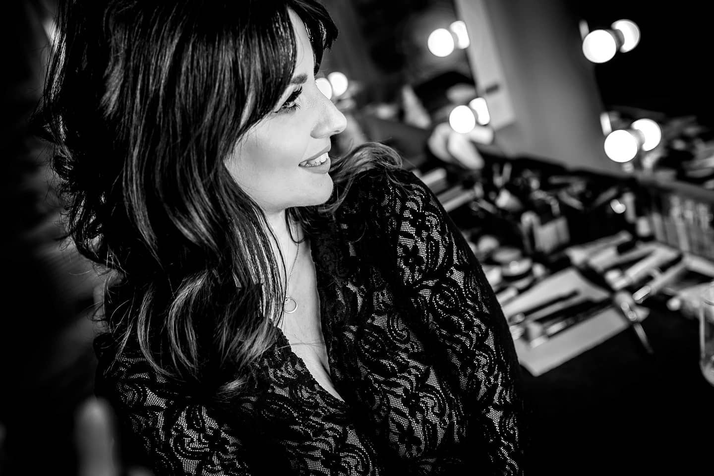 Portret kobiety z profilu, uśmiechniętej, w bluzce z koronki, z rozmazanym tłem stanowiska do makijażu z lampami.