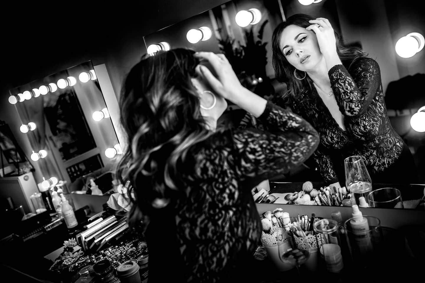 Czarno-białe zdjęcie kobiety w koronkowej bluzce, poprawiającej włosy przed lustrem z oświetleniem, otoczonej kosmetykami i akcesoriami do makijażu.