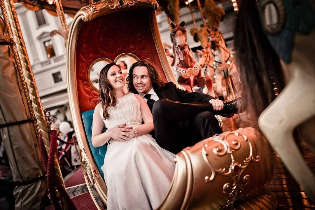 Uśmiechnięta para narzeczonych siedzi w złotej karocy na karuzeli, mężczyzna w czarnym garniturze i kobieta w białej sukni, w tle konie karuzeli i budynki miejskie.