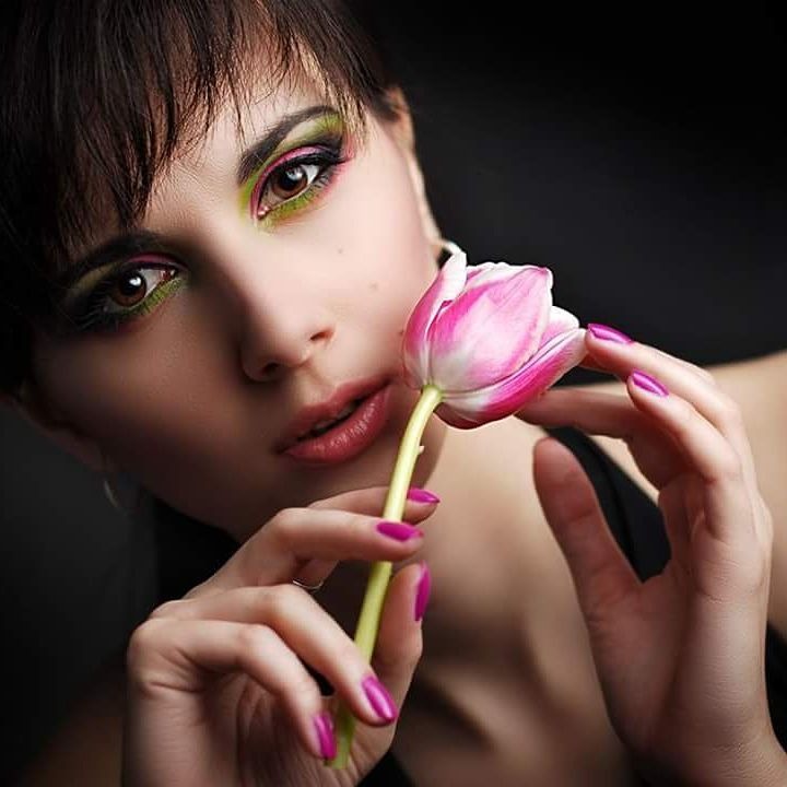 Portret kobiety z intensywnym makijażem oczu w odcieniach zieleni i różu, trzymającej różowego tulipana przy twarzy, z paznokciami pomalowanymi na fuksjowy kolor.