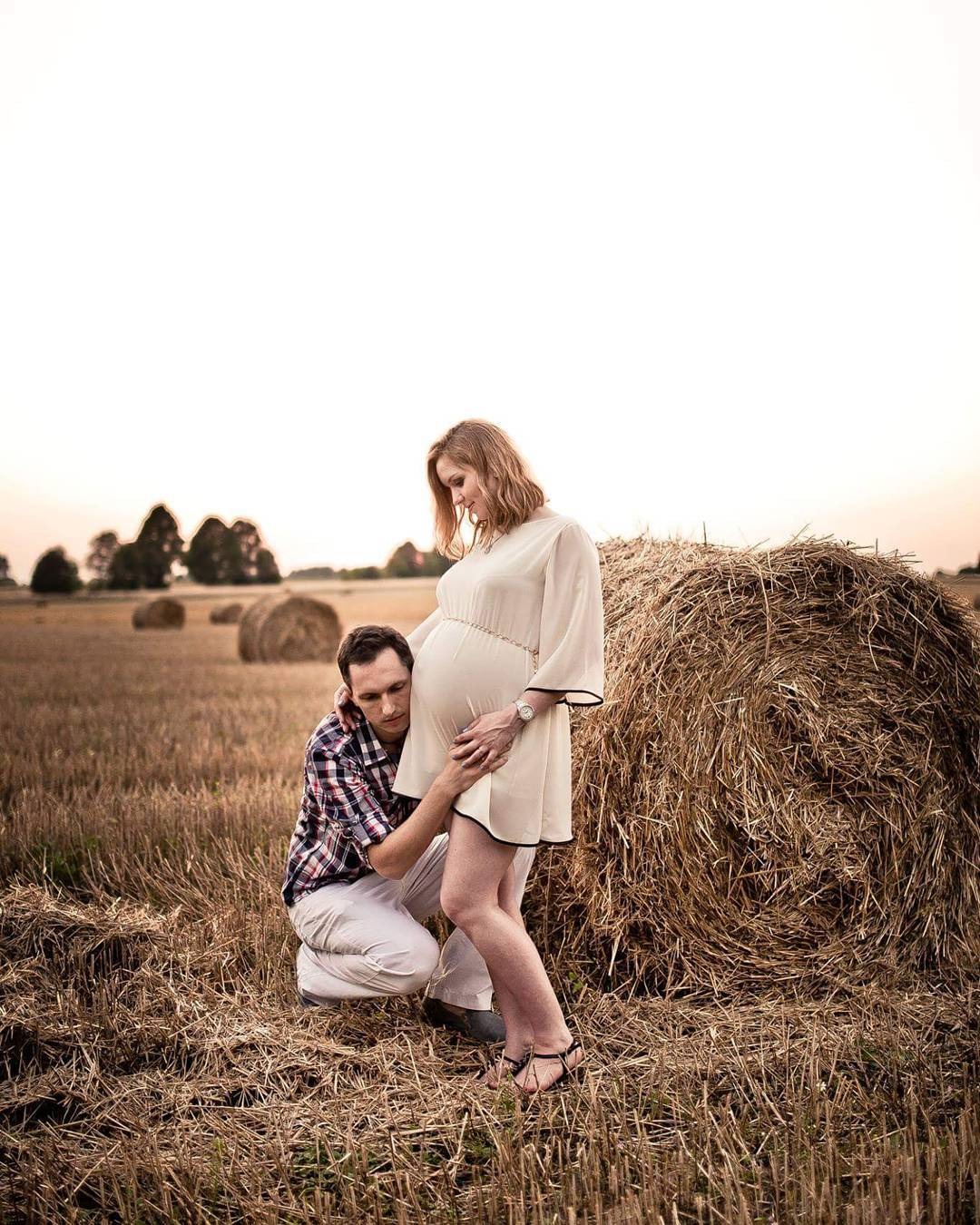 Przyszły tata słucha brzucha ciężarnej partnerki na tle pola ze słomianymi balotami, w ciepłym świetle zachodzącego słońca.