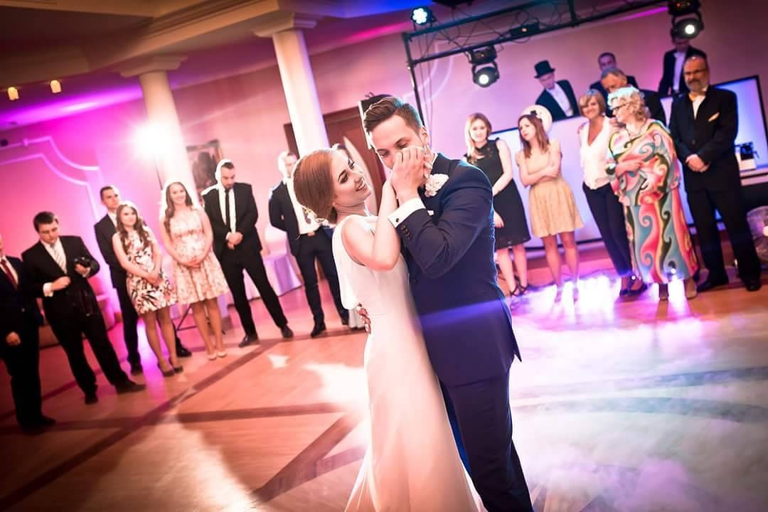 Para młoda w tańcu podczas wesela, panna młoda w prostej białej sukni, pan młody w granatowym garniturze, goście weselni w tle.