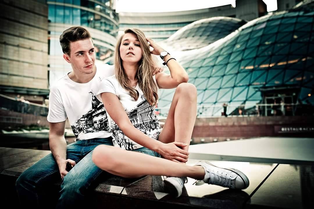 Para młodych ludzi w miejskiej scenerii, ona siedzi na murku w białych trampkach i koszulce z nadrukiem miasta, on siedzi obok w jeansach i koszulce z podobnym motywem, na tle nowoczesna architektura.