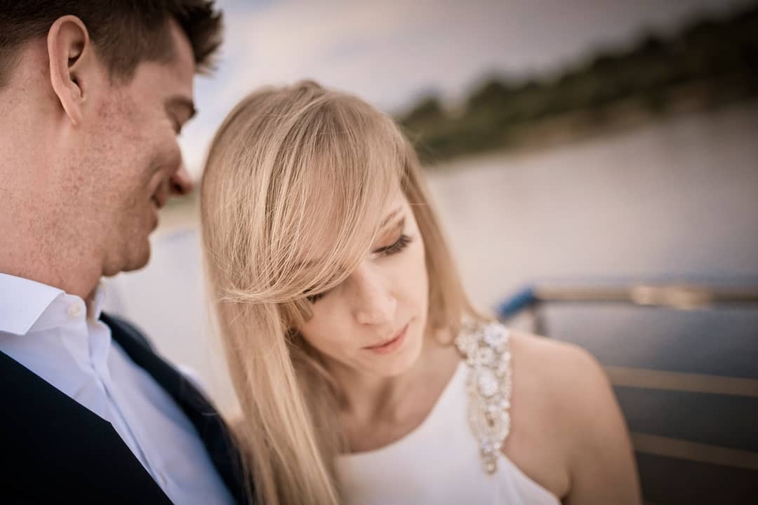 Para narzeczonych: blond kobieta w białej sukience z ozdobnym ramieniem spogląda w dół, obok mężczyzna w czarnej kamizelce i białej koszuli, tło rozmyte, widoczny fragment rzeki lub jeziora.