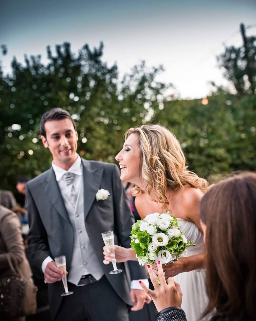 Uśmiechnięta panna młoda w białej sukni trzyma bukiet białych róż i szampana, obok pan młody w szarym garniturze z kieliszkiem szampana, rozmawiają z gościem weselnym