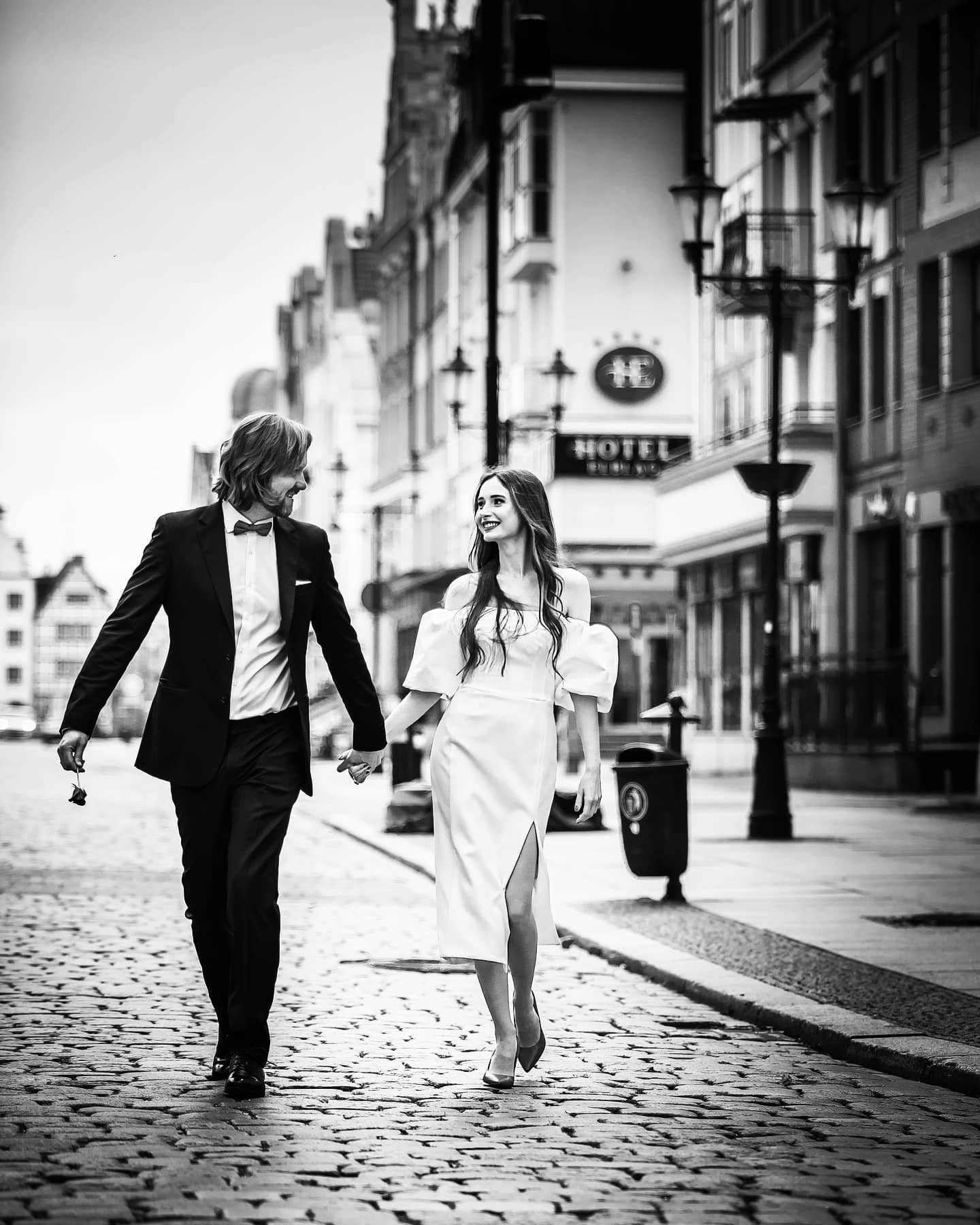 Czarno-białe zdjęcie pary młodej spacerującej po brukowanej ulicy miejskiej; mężczyzna w smokingu trzyma zwiędłą różę, kobieta w białej sukni z rozcięciem uśmiecha się, w tle budynki i latarnie.