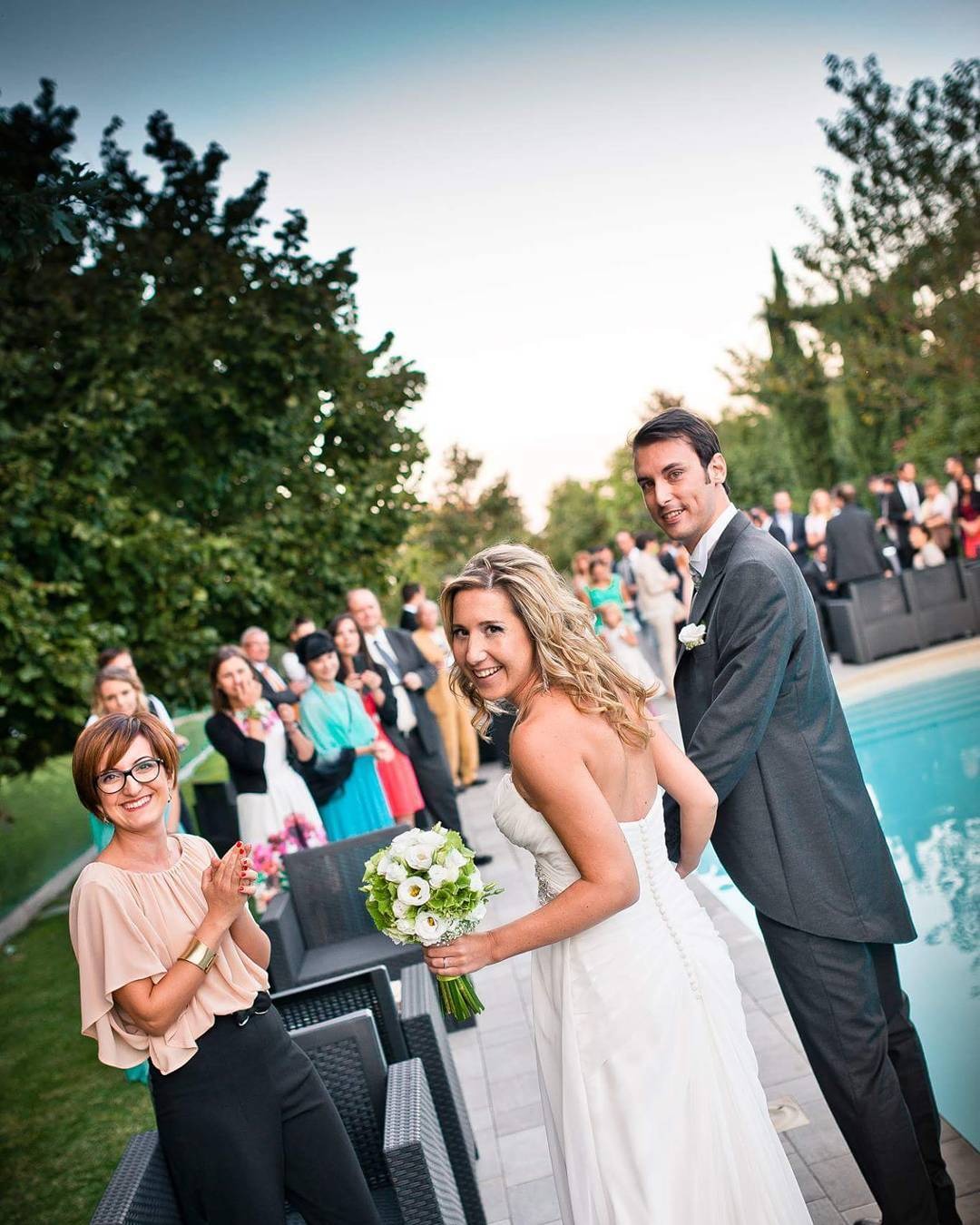 Uśmiechnięta panna młoda w białej sukni i pan młody w szarym garniturze, trzymający się za ręce, otoczeni gośćmi weselnymi i basenem w tle, kobieta w okularach klaszcze w dłonie.