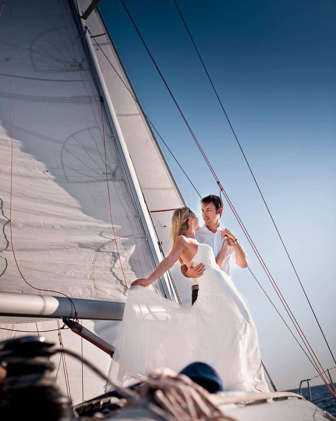 Para młoda w białych strojach pozuje na jachcie z rozpiętymi żaglami na tle błękitnego nieba, w tle widoczne liny i elementy wyposażenia łodzi.