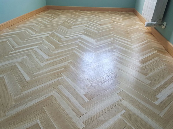 Jasny parkiet ułożony we wzór jodełki francuskiej, widoczny grzejnik przy ścianie i listwa przypodłogowa.