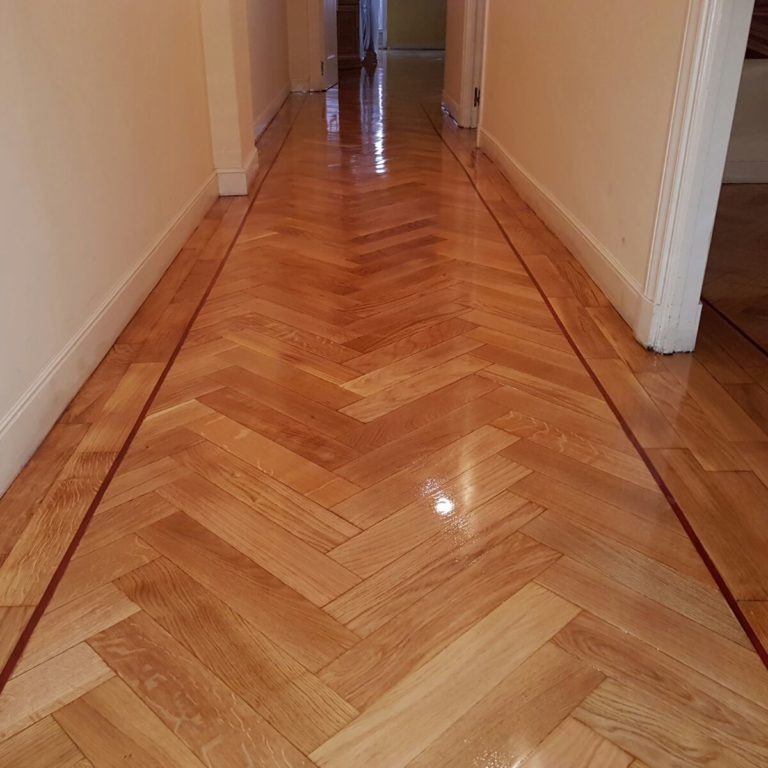 Polerowana podłoga parkietowa ułożona we wzór jodełki w korytarzu, z ciemniejszą bordiurą wzdłuż ścian i błyszczącą powierzchnią odbijającą światło.