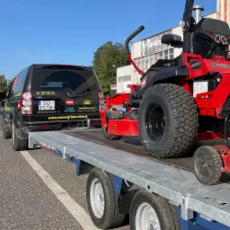 Czerwona kosiarka samojezdna Toro 4000 Pro na przyczepie za czarnym Land Roverem Discovery 4 z naklejkami firmy ogrodniczej Snow Grass Expert, w tle budynek z napisem Bawialnia i błękitne niebo.