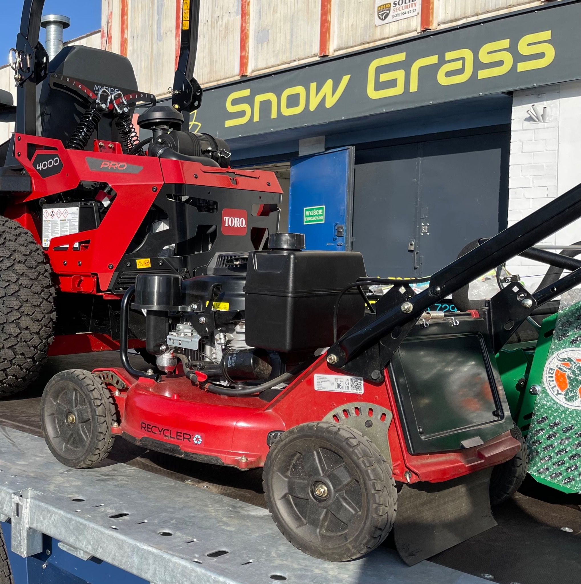 Czerwona kosiarka samojezdna Toro Recycler z serii 4000 PRO, zaparkowana na metalowej platformie transportowej, z widocznym logo 'Snow Grass' w tle.