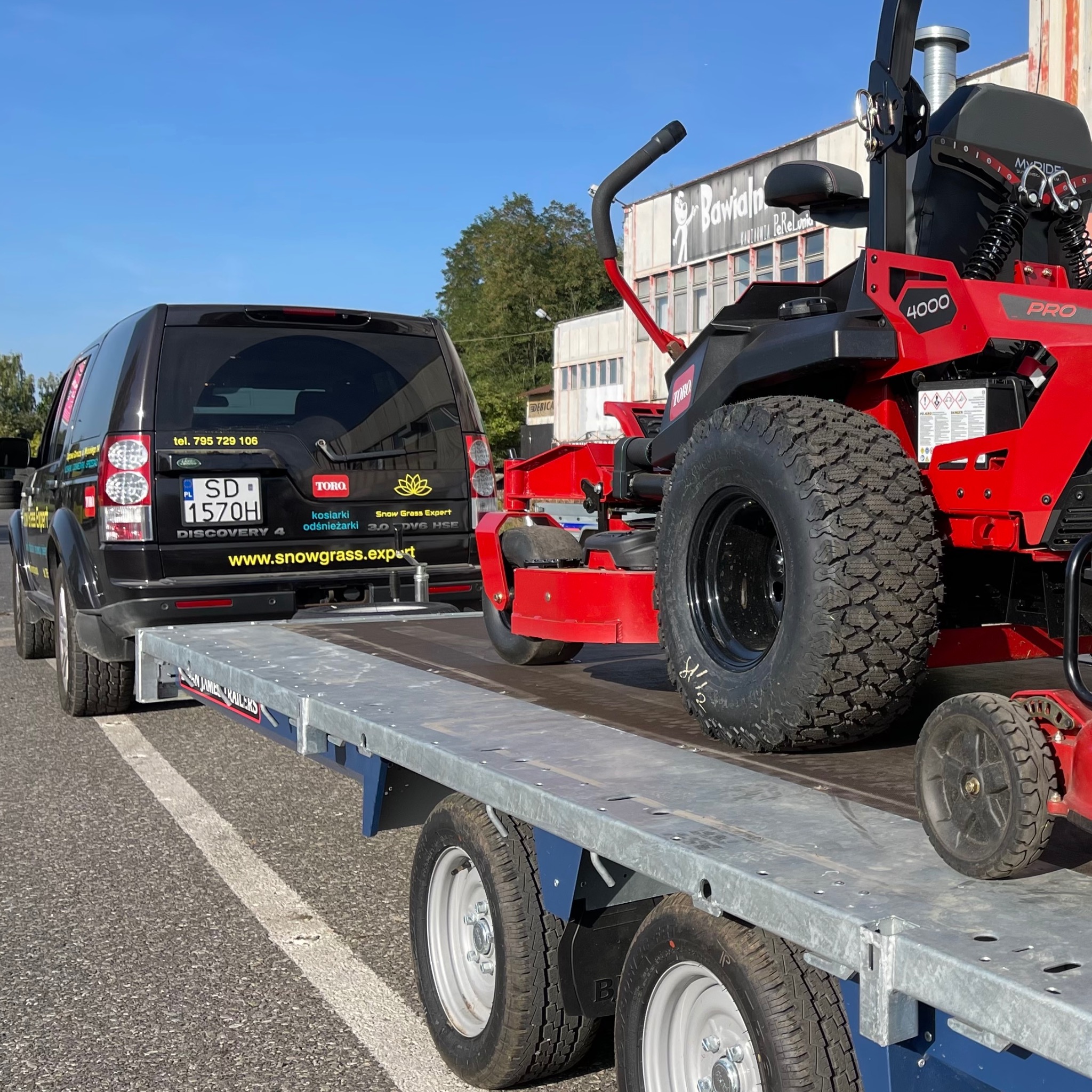 Czerwona kosiarka samojezdna Toro 4000 Pro na przyczepie za czarnym Land Roverem Discovery 4 z naklejkami firmy ogrodniczej Snow Grass Expert, w tle budynek z napisem Bawialnia i błękitne niebo.