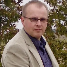 Portret mężczyzny w okularach i beżowej marynarce na tle zielonych krzewów.