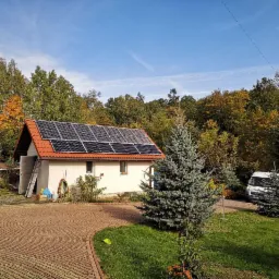 Biały dom jednorodzinny z czerwonym dachem i panelami słonecznymi, otoczony zielenią i brukowanym podjazdem, z widoczną drabiną opartą o ścianę i samochodem dostawczym w tle.